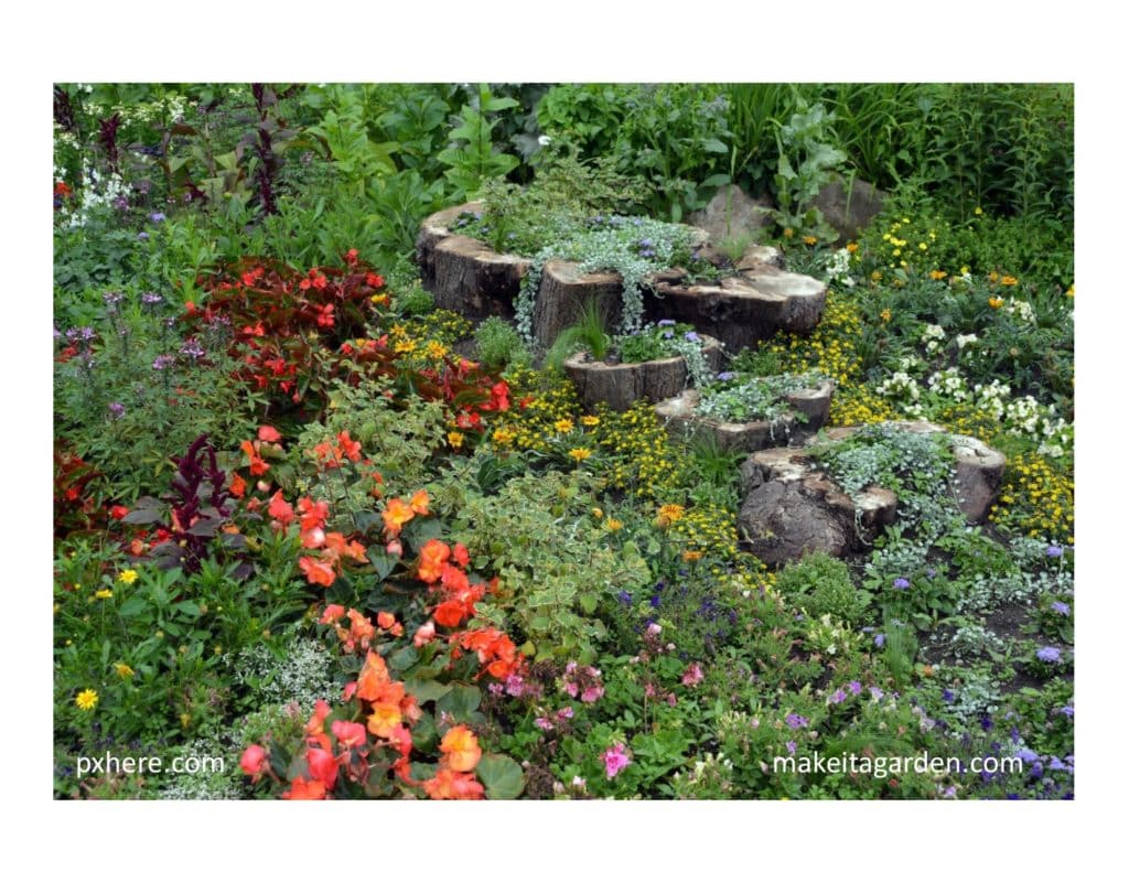 garden design trends of 2020 picture of a flower bed filled with blooming annual and perennials. There are short tree stumps  with trailing vine plants planted in the center. The stumps make garden look natural and woodsy.