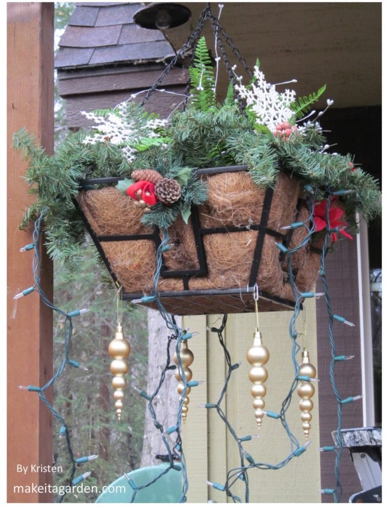 For an Elegant Holiday Porch, make a hanging basket lush with Christmas greenery.  Photo shows close up of basket with artificial pine branches, fern, pine cones, holly and snowflake 