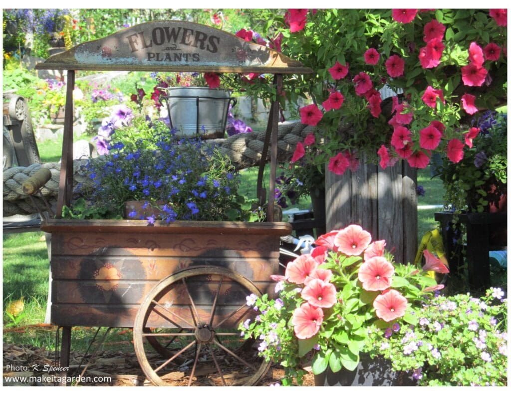 dazzling flower garden. Large wooden cart with wooden wheel. Flowers inside spilling over the edge and baskets of pink flowers next to it. 