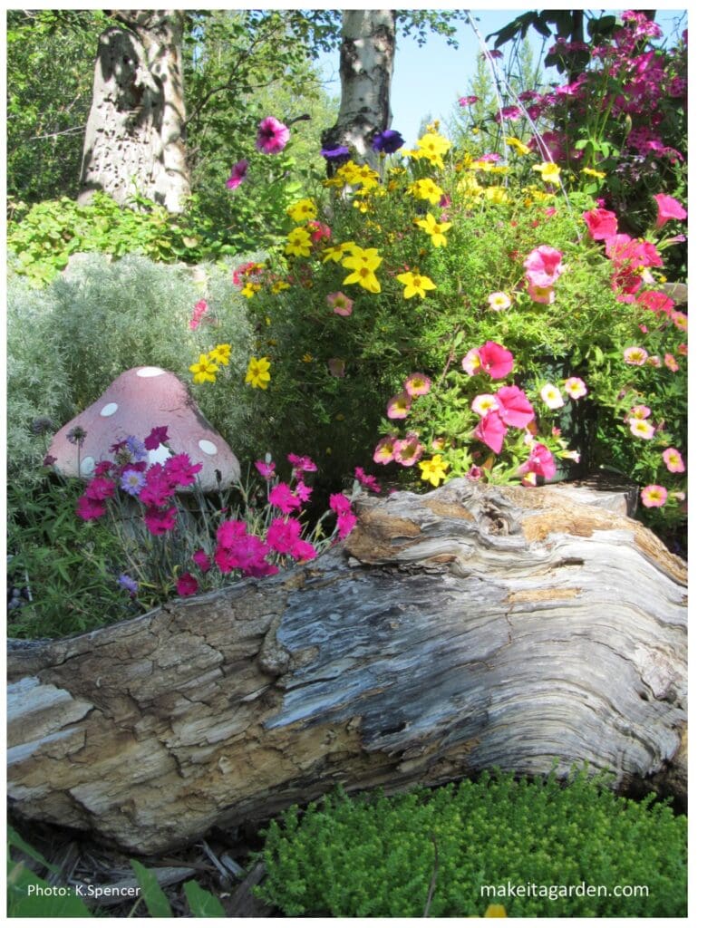 Large but short piece of an old tree trunk is weathered and rotting. Surrounded by flowers and ceramic mushroom garden ornament.