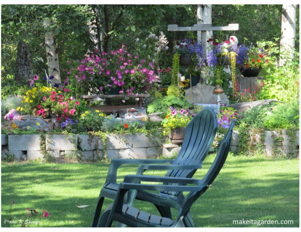 2 easy chairs on a lawn with lots of colorful flowers in background. Driftwood makes a happy garden