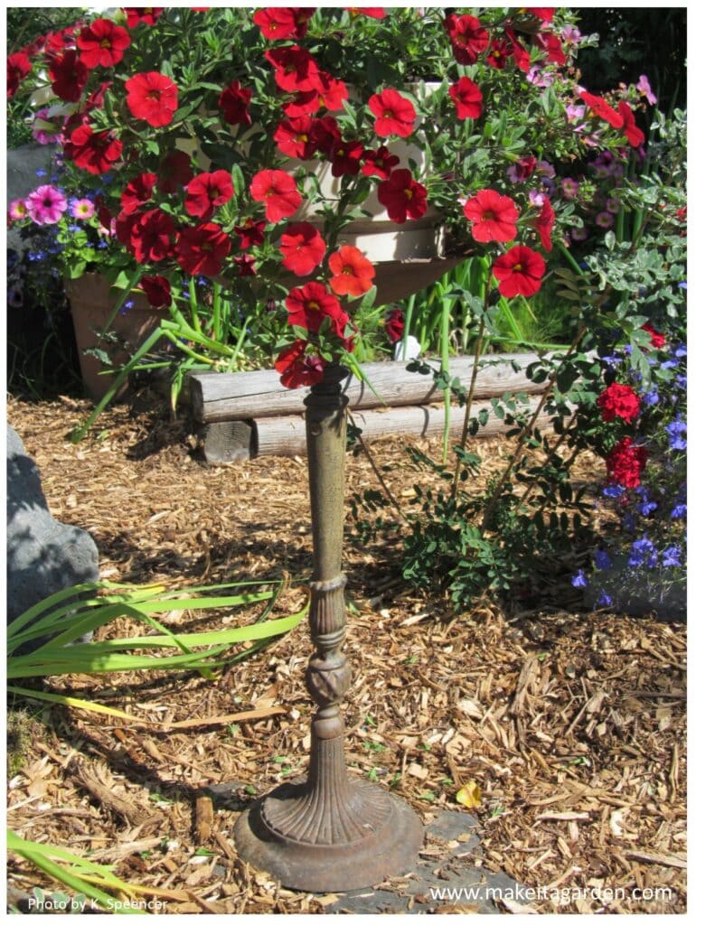 Close up of an antique metal bird bath that looks like a tall lamp stand. It is used to display basket of flowers on top.
