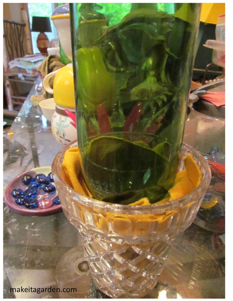 Wine bottle upside down in a glass vase with cloth napkin tucked inside to hole the bottle steady