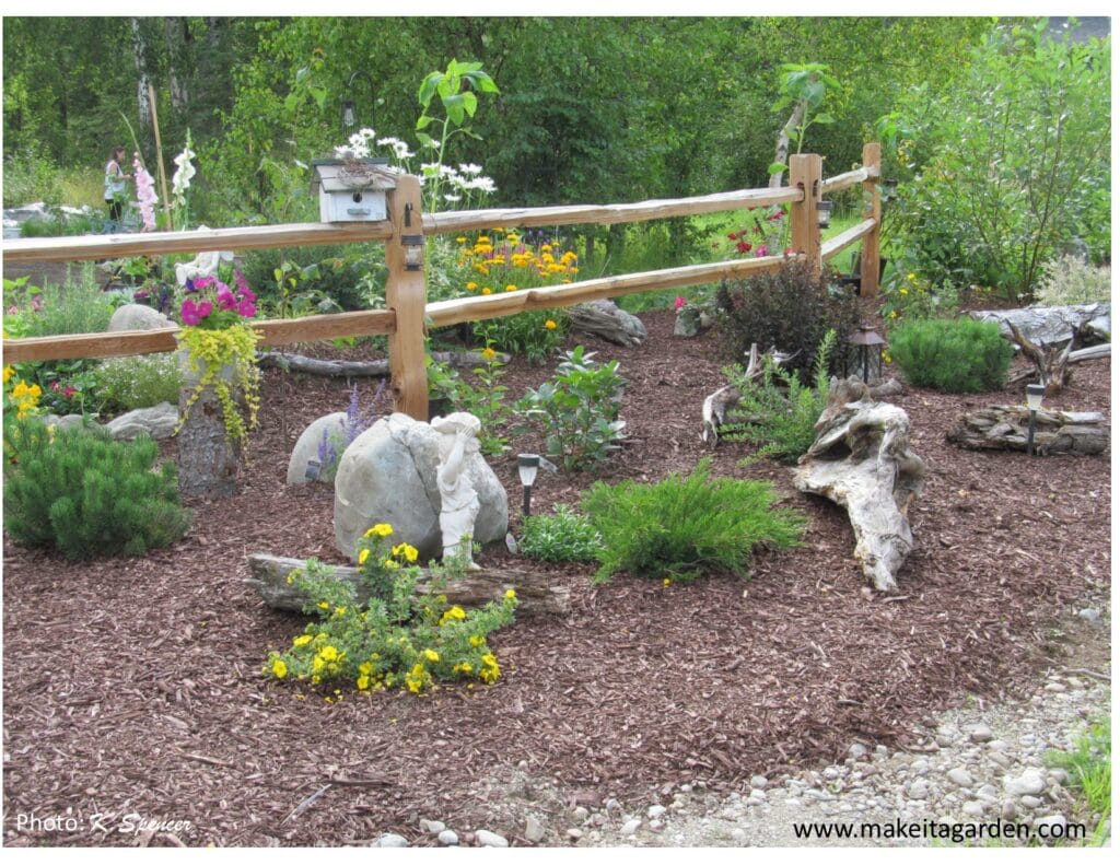 Wooden split rail fence with rocks and flowers and pieces of driftwood in front of it and behind it. A start to a spectacular flower garden
