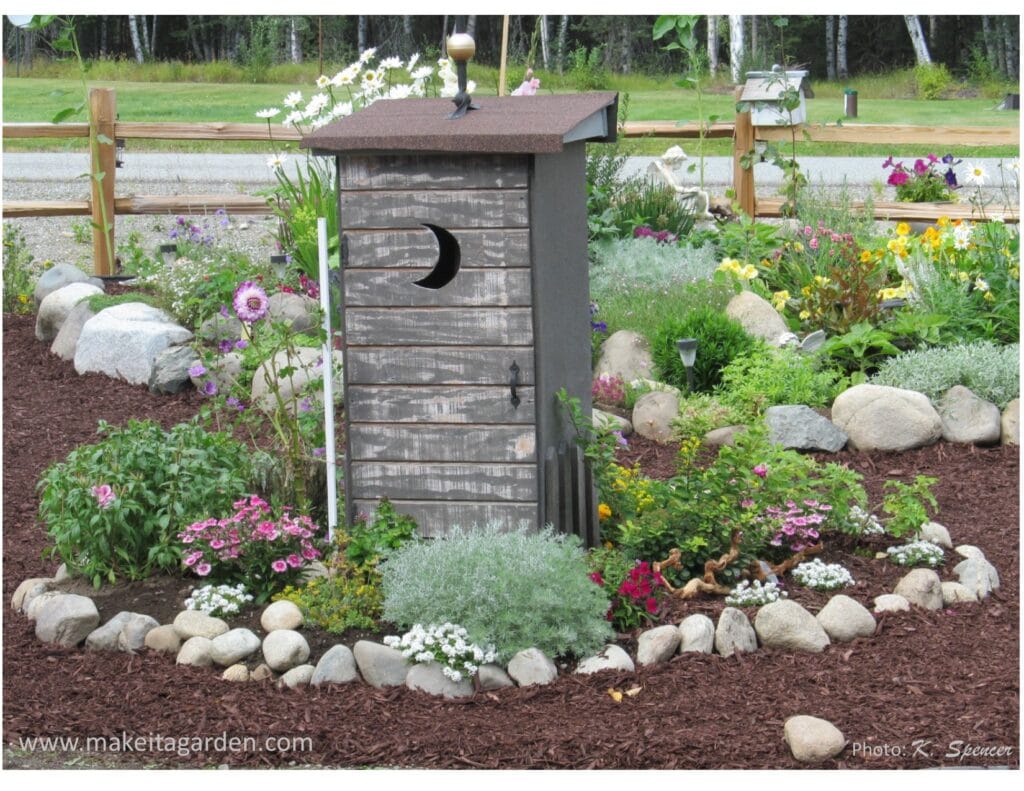 Decorative "fake" outhouse as garden decor. Surrounded by numerous blooming flowers in the rock garden