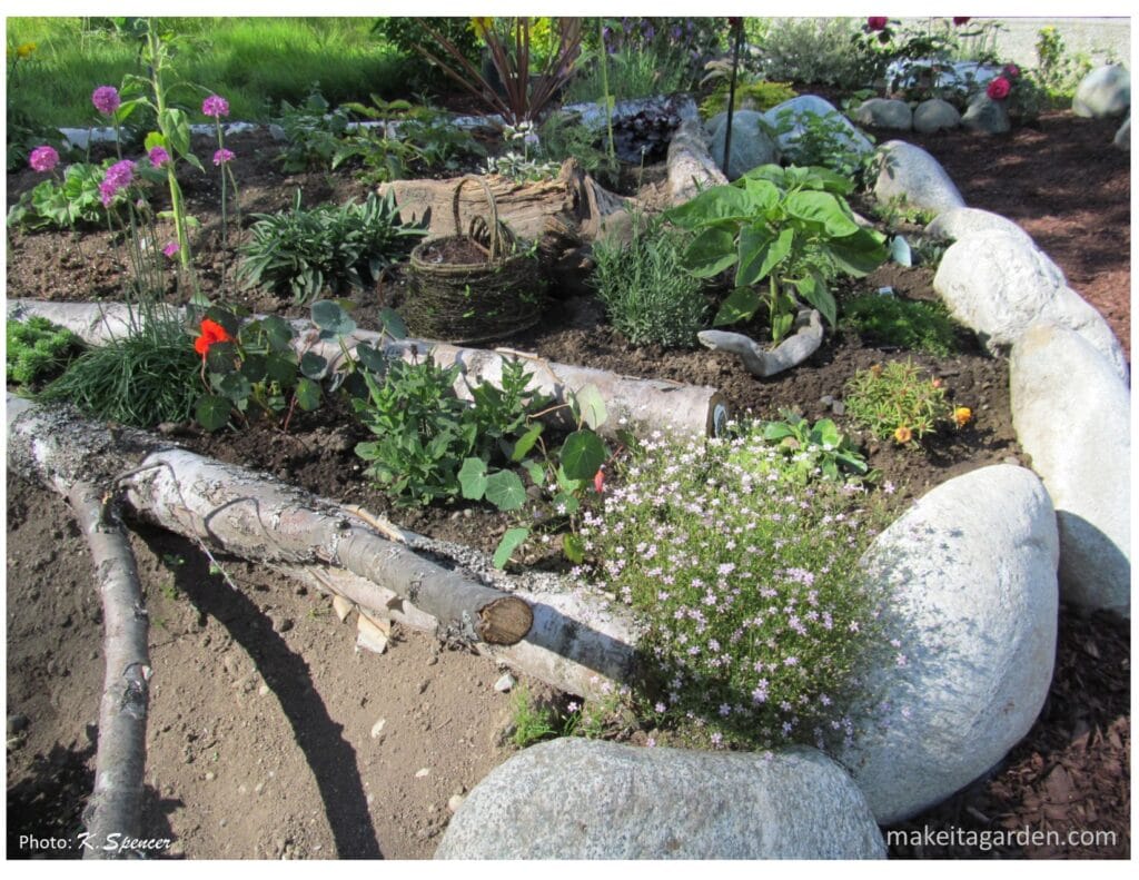 garden bed lined with boulders and a small diameter log lying across the dirt with flowers planted around it