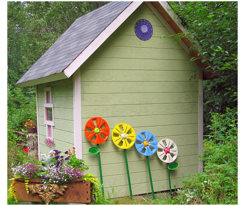 4 brightly painted hub cap flowers lean against the garden shed next to real flowers